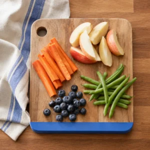 Wooden cutting board with blue painted edge holding sliced apple, carrot sticks, blueberries and green beans in soft daylight