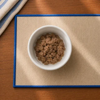 Small white bowl with novel-protein dog food on cream placemat with blue trim, top-down