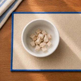 White ceramic bowl with small portion of plain chicken on cream placemat with blue trim